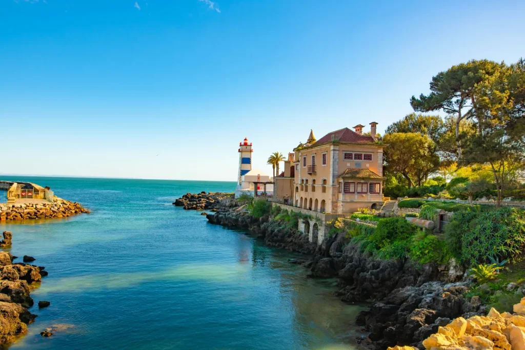 A historic building and a blue-and-white-striped lighthouse on the coast of Cascais, Portugal.